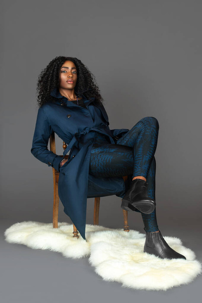 Portrait of a young black woman with long curly black hair, beautiful makeup sitting by herself on fur in a studio with a grey background wearing a blue trench coat and leggings with black boots. - Photo, Image