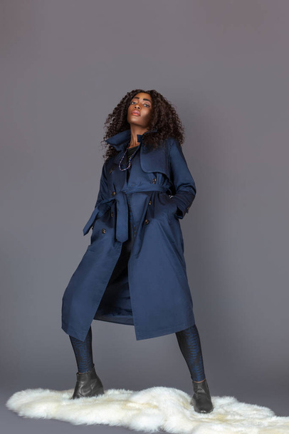 Portrait of a tall black woman with long curly black hair, beautiful makeup posing by herself on fur in a studio with a grey background wearing a blue trench coat and leggings with black boots. - Photo, image