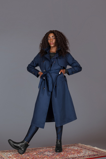 Portrait of a tall black woman with long curly black hair, beautiful makeup posing by herself on a carpet in a studio with a grey background wearing a blue trench coat and leggings with black boots. - Fotografie, Obrázek