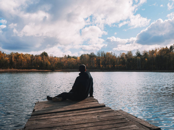 Młody człowiek na drewnianym moście nad jeziorem, w ponury jesienny dzień. Atmosfera samotności i smutku - Zdjęcie, obraz