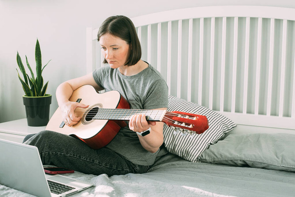 Egy fiatal nő videóleckét néz a gitározásról otthon. Online gitárórák. A hobbi koncepciója, szabadidős tevékenységek a karantén alatt, önelkülönítés. - Fotó, kép