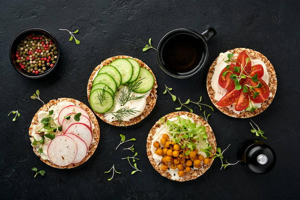 Four crispy buckwheat bread with cream cheese, radish, tomato, chickpea, cucumber and microgreen for healthy breakfast on parchment paper on black stone background. Concept vegan and healthy eating. - Photo, image