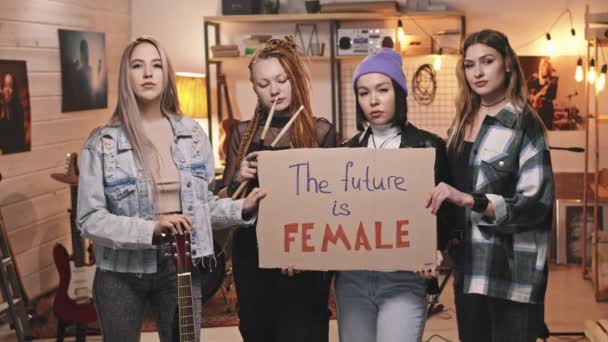 Medium slowmo portrait of cool all-girl rock band looking at camera confidently holding cardboard sign saying future is female. Концепция расширения прав и возможностей женщин - Кадры, видео