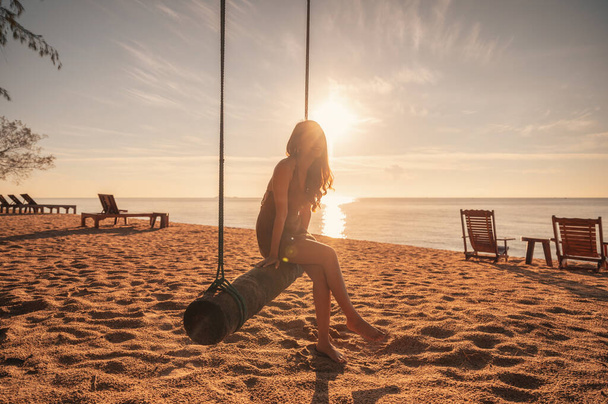 若いアジアの女性は熱帯の海で日の出のビーチで木製のスイングでリラックス - 写真・画像