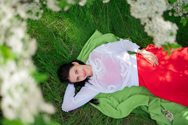 A young woman lies on a plaid on the lawn under the branches of a flowering tree - Fotografie, Obrázek