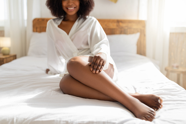 Vue recadrée de la jeune femme noire assise sur le lit, portant un peignoir, touchant sa jambe, jouissant d'une peau lisse après l'épilation - Photo, image