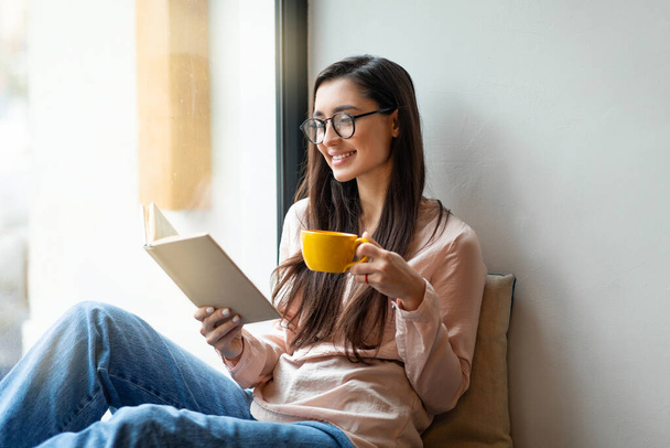 Після карантину. Щаслива арабська жінка читає книгу і насолоджується ранковою кавою, відпочиває в кафе біля вікна
 - Фото, зображення