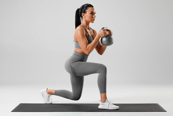 Active girl doing lunges exercises with kettlebell, leg muscle training. Fitness woman doing front forward one leg step lunge - 写真・画像