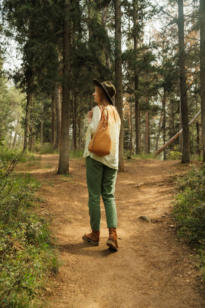 Achteraanzicht van vrouw met rugzak en hoed wandelen in bos - Foto, afbeelding