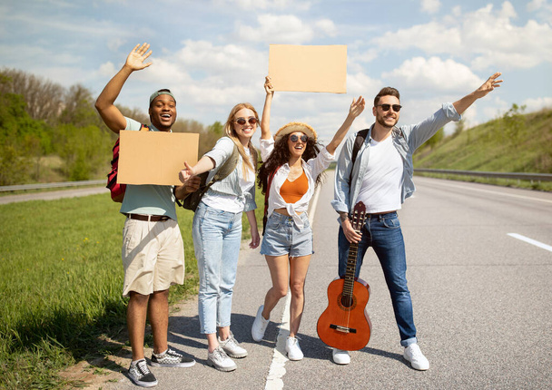 Glückliche Freunde stehen mit leerem Schild auf der Autobahn, winken Auto herunter, gehen gemeinsam auf Reisen, draußen - Foto, Bild