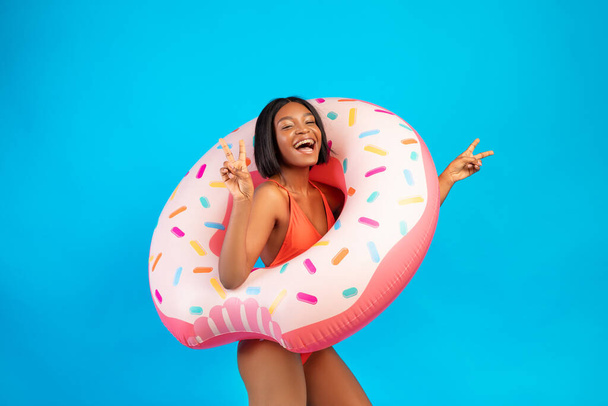 Unbekümmerte schwarze Frau im Badeanzug trägt aufblasbaren Donut-Ring, lacht und macht Friedensgeste auf blauem Hintergrund - Foto, Bild