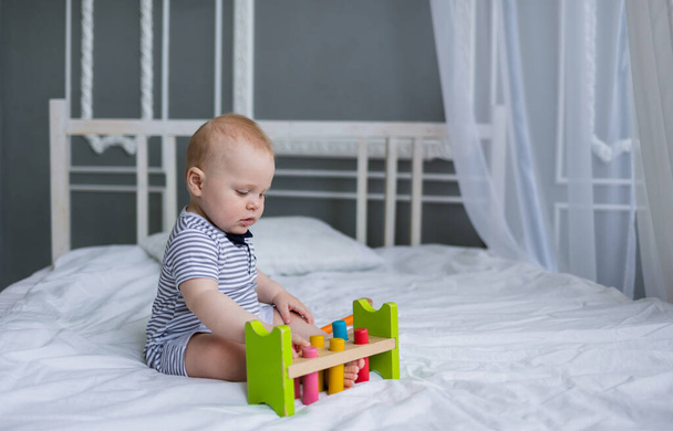 bebé niño se sienta en un mono a rayas y juega con un juguete de madera en una cama blanca en la habitación - Foto, Imagen
