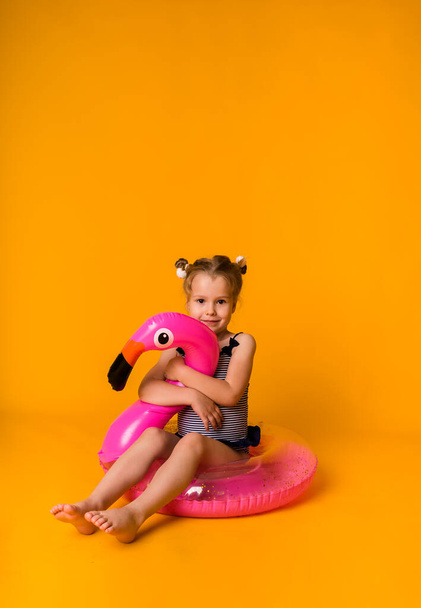 Dziewczynka w pasiastym stroju kąpielowym siedzi na różowym nadmuchiwanym okręgu z flamingiem na żółtym tle z miejscem na tekst. letnie wakacje z dzieckiem - Zdjęcie, obraz