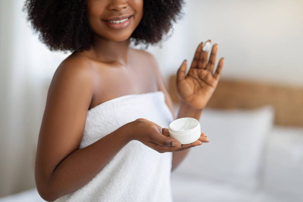 Attractive young black female in bath towel applying natural moisturizing cream on her face at home, closeup view - Valokuva, kuva