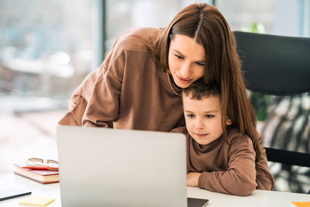 Liefdevolle moeder helpt zoon met huiswerk terwijl hij achter de laptop zit. Vrouw met een jongetje achter de computer - Foto, afbeelding