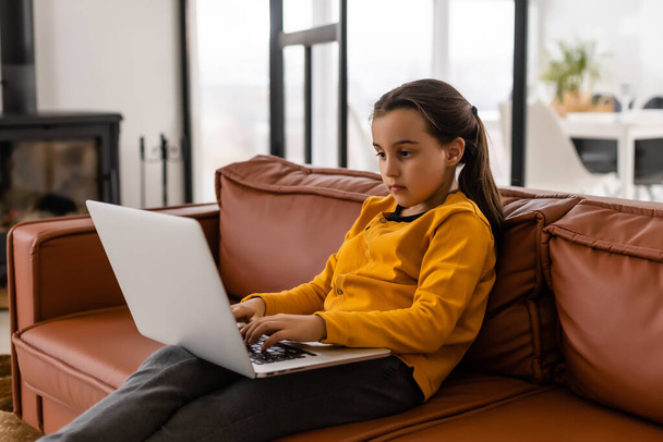 Smart Little Girl Does Homework in Her Living Room. Shes Sitting and Uses Laptop. - Photo, image