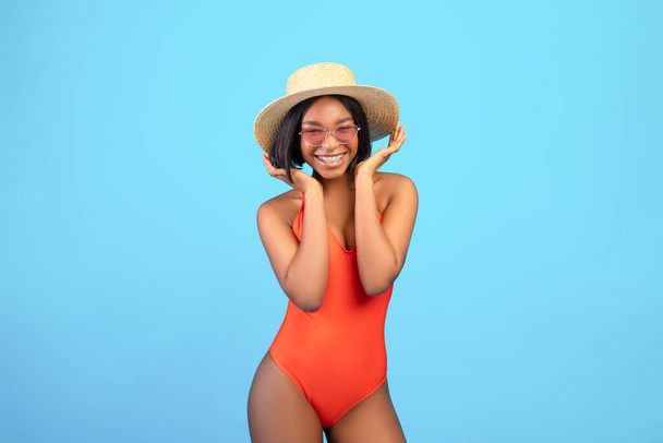 Ritratto di donna nera spensierata in bikini, occhiali da sole e cappello di paglia in posa e sorridente davanti alla macchina fotografica su sfondo blu - Foto, immagini