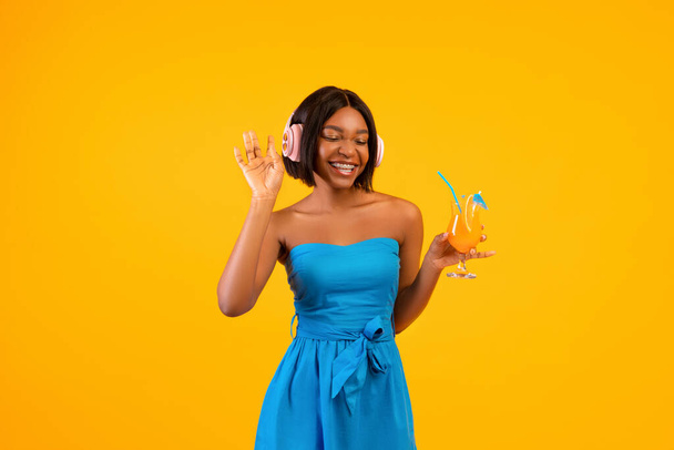 Zwarte dame in stijlvolle outfit met exotische cocktail en genieten van favoriete muziek in koptelefoon op oranje achtergrond - Foto, afbeelding