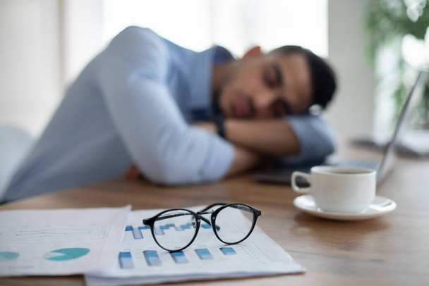 Hombre de negocios árabe agotado durmiendo en su escritorio de la oficina, cansado de exceso de trabajo, enfoque selectivo en documentos y gafas - Foto, Imagen