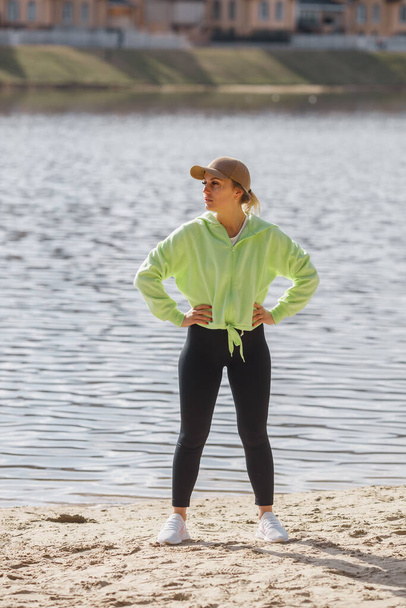 Veautiful self determied female runner in trendy sportswear posing isolated on river bank, stretching legs, warming up muscles before running exercise outdoors early in the morning. - Photo, Image