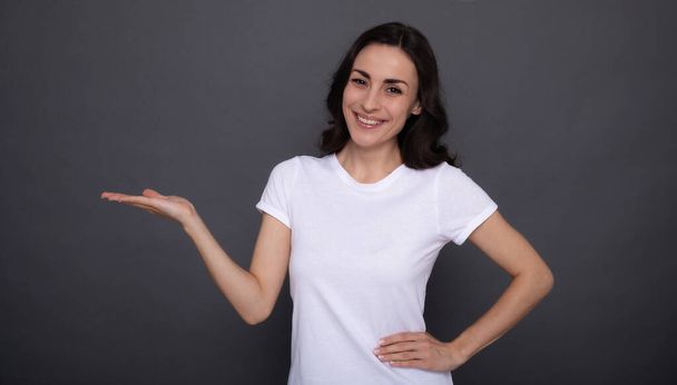 Young beautiful stylish happy woman in a white t-shirt is posing and pointing away on the gray background - 写真・画像