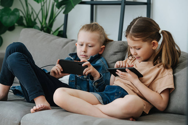 Little siblings sitting on the couch, watching their phones next to each other - Foto, Imagen