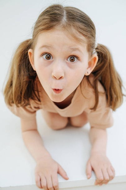 Funny little girl with beautiful hazel eyes standing on all fours, grimacing over white backgound. Making surprised face and wide eyes. Face close to camera. - 写真・画像