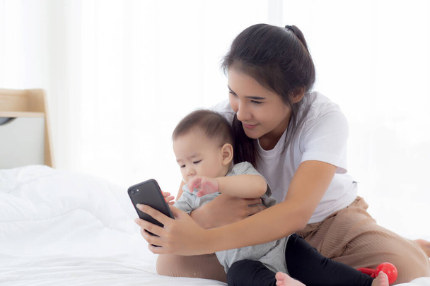 Jonge Aziatische moeder en kleine baby meisje of pasgeboren selfie met smartphone op bed in de slaapkamer, geluk mam en dochter met behulp van telefoon video gesprek thuis, twee mensen, familie en communicatie concept. - Foto, afbeelding