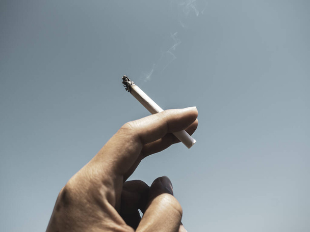 Cigarette in man hand with smoke on blue sky background. Hand holding cigarette. - 写真・画像