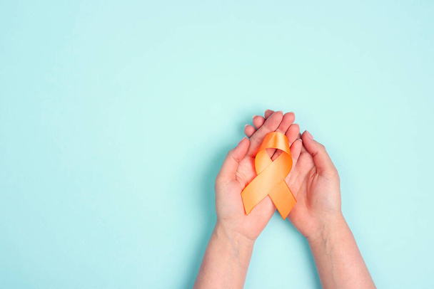 Weibliche Hände halten eine orangefarbene Schleife auf blauem Hintergrund. Welttag der Multiplen Sklerose. Kopierraum für Text. - Foto, Bild
