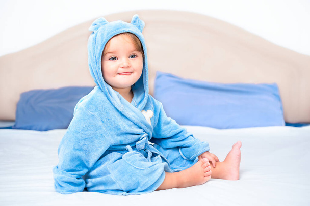 Ein Kind im Mantel sitzt auf dem Bett. Niedliches Baby lächelt nach dem Baden - Foto, Bild