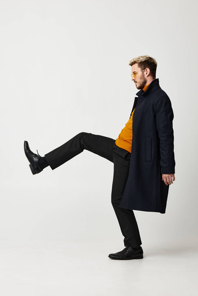 energetic guy raised his leg up on a light background and a classic coat orange sweater pants - Foto, imagen