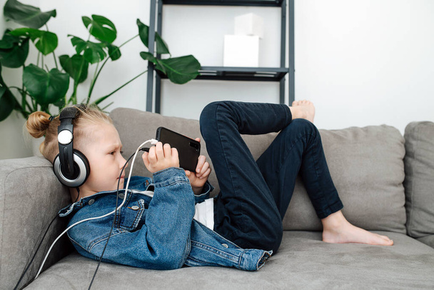 Konzentrierter Junge mit Kopfhörern, der sein Handy auf einer Couch betrachtet und im Wohnzimmer Spiele spielt. Seitenansicht. - Foto, Bild