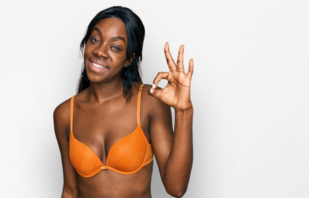 Genç Afro-Amerikalı kadın iç çamaşırı giyiyor, gülümsüyor ve el ve parmaklarıyla "Tamam" işareti yapıyor. başarılı ifade.  - Fotoğraf, Görsel