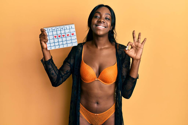 Молодая африканская американка в нижнем белье держит календарь сердца делает хорошо знак с пальцами, улыбаясь дружелюбный жест отличный символ  - Фото, изображение