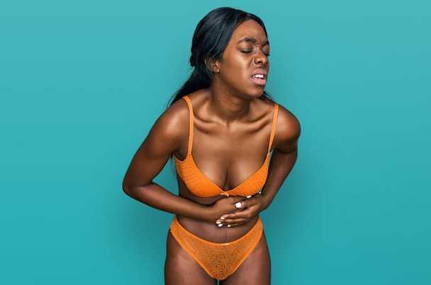 吐き気、痛みを伴う病気が具合が悪いので、若いアフリカ系アメリカ人女性は胃の上に手でランジェリーを着ています。キャッシュ・コンセプト.  - 写真・画像