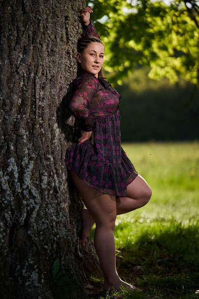 Plus size beautiful woman in summer dress by a huge oak in the forest - Фото, изображение