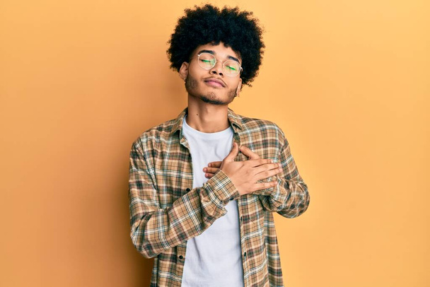 Jonge Afro-Amerikaanse man met afrohaar in casual kleding glimlachend met handen op de borst, ogen dicht met dankbaar gebaar op gezicht. Gezondheidsconcept.  - Foto, afbeelding