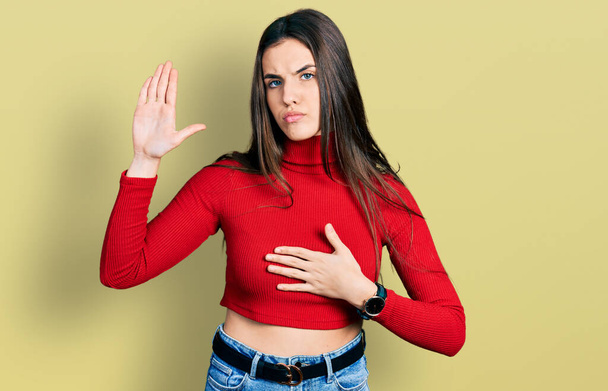 Giovane adolescente bruna indossa maglione rosso collo alto giurando con mano sul petto e palmo aperto, facendo un giuramento di fedeltà  - Foto, immagini