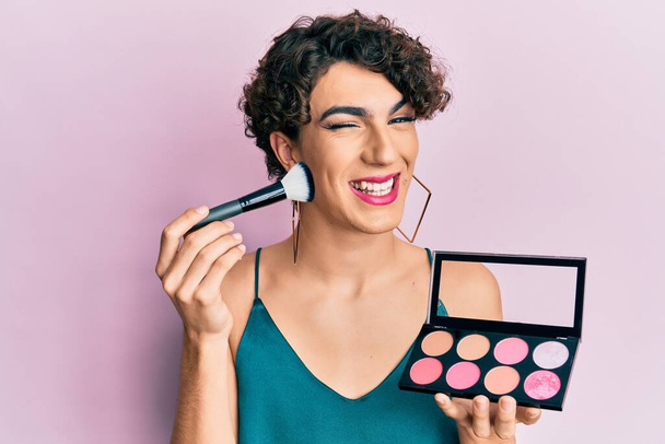 Молодой человек в женском макияже, держа кисть для макияжа и краснея подмигивая, смотрит в камеру с сексуальным выражением лица, веселым и счастливым лицом.  - Фото, изображение