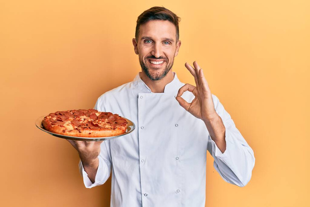 Handsome man with beard professional cook holding italian pizza doing ok sign with fingers, smiling friendly gesturing excellent symbol  - Foto, afbeelding