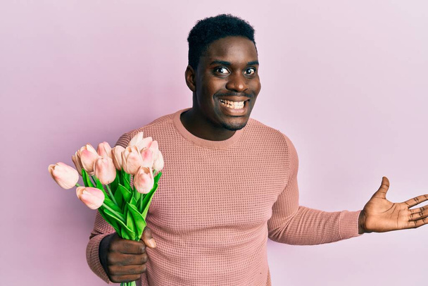 Przystojny czarny człowiek gospodarstwa bukiet różowe tulipany kwiaty świętując zwycięstwo z uśmiechem szczęśliwy i zwycięzca ekspresji z podniesionymi rękami  - Zdjęcie, obraz