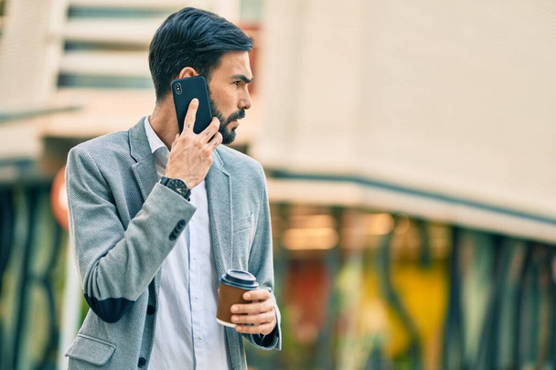 Młody latynoski biznesmen z poważnym wyrazem twarzy rozmawiający przez smartfona i pijący kawę w mieście. - Zdjęcie, obraz