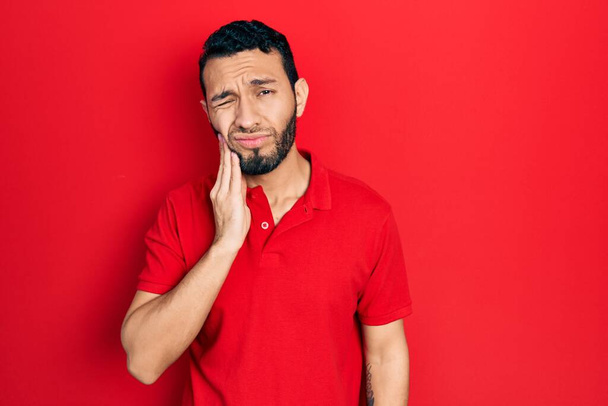 Hombre hispano con barba con camiseta roja casual tocando la boca con la mano con expresión dolorosa debido a dolor de muelas o enfermedad dental en los dientes. dentista  - Foto, imagen