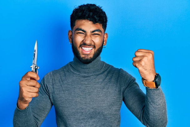Arab man with beard holding pocket knife screaming proud, celebrating victory and success very excited with raised arms  - Photo, Image