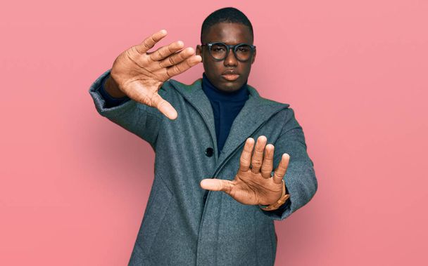 Giovane uomo afroamericano che indossa abiti da lavoro e occhiali facendo cornice con le mani palme e le dita, prospettiva fotocamera  - Foto, immagini