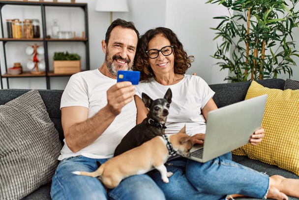 Latynoska para w średnim wieku, używająca laptopa i karty kredytowej. Siedzenie na kanapie z psami w domu. - Zdjęcie, obraz