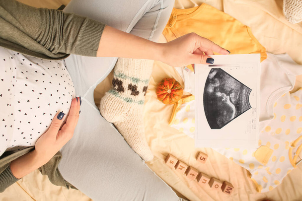 Femme enceinte avec image d'échographie et accessoires bébé sur le lit, gros plan - Photo, image