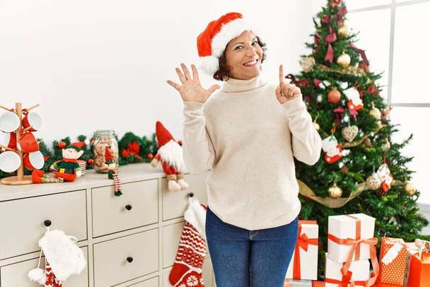Mujer hispana de mediana edad de pie junto al árbol de navidad mostrando y señalando hacia arriba con los dedos número seis mientras sonríe confiada y feliz.  - Foto, imagen