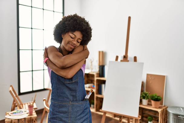 Młoda Afroamerykanka z afro włosami w studiu artystycznym przytulająca się szczęśliwa i pozytywna, uśmiechnięta pewna siebie. miłość własna i opieka własna  - Zdjęcie, obraz